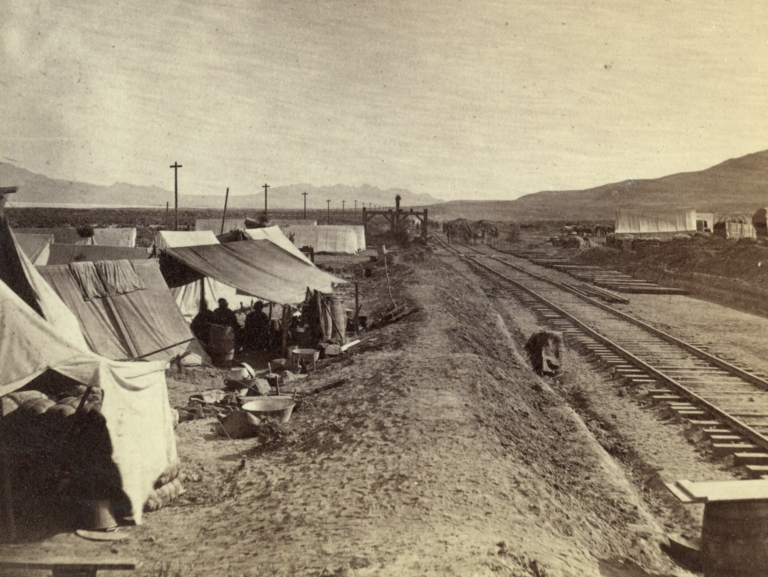 Black and white photography of a camp set up parallel to a railroad. Tents and tools are visible. A group of men sit under a tent.